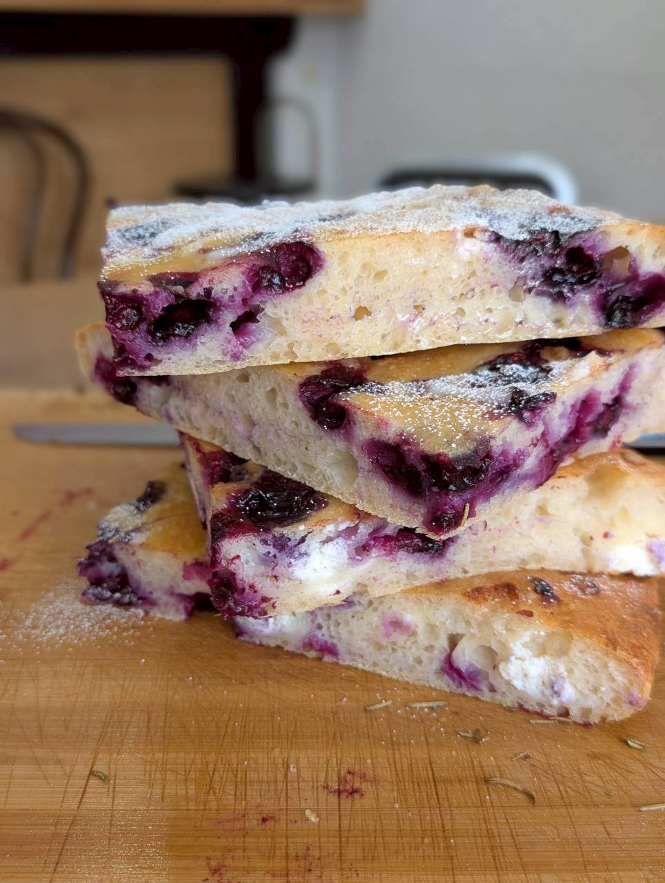Three stacked slices of blueberry focaccia on a wooden board, showing the purple-stained open crumb and powdered sugar dusting — Focaccino Hakuba