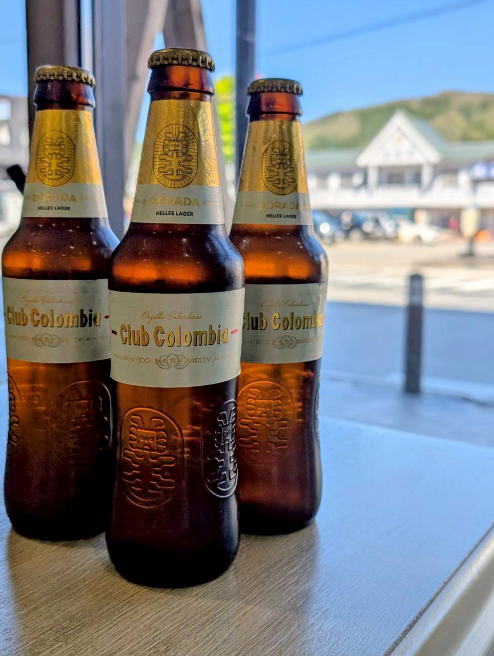 Three Club Colombia Dorada lager bottles on a table with Hakuba Station in the background — Focaccino Hakuba