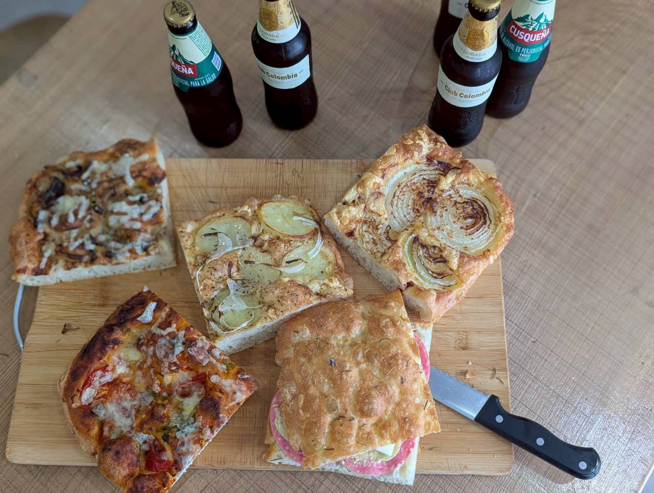 Top-down selection of five focaccia squares with Colombian and Peruvian craft beers and a knife on a wooden board — Focaccino Hakuba